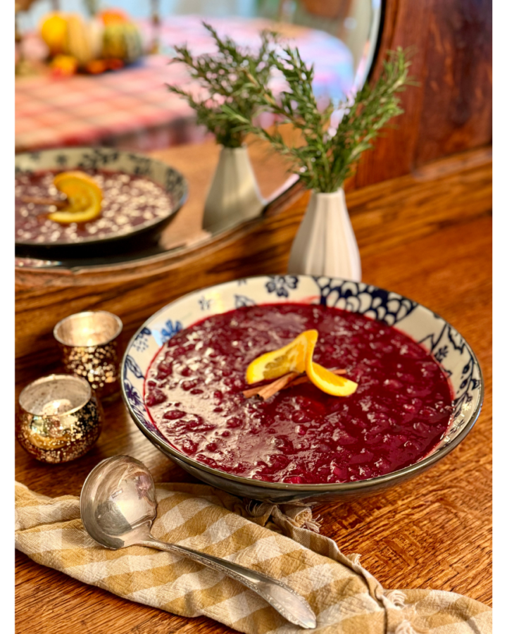 Bowl of cranberry sauce on wooden hutch with vase of rosemary and gingham cloth napkin and serving spoon.