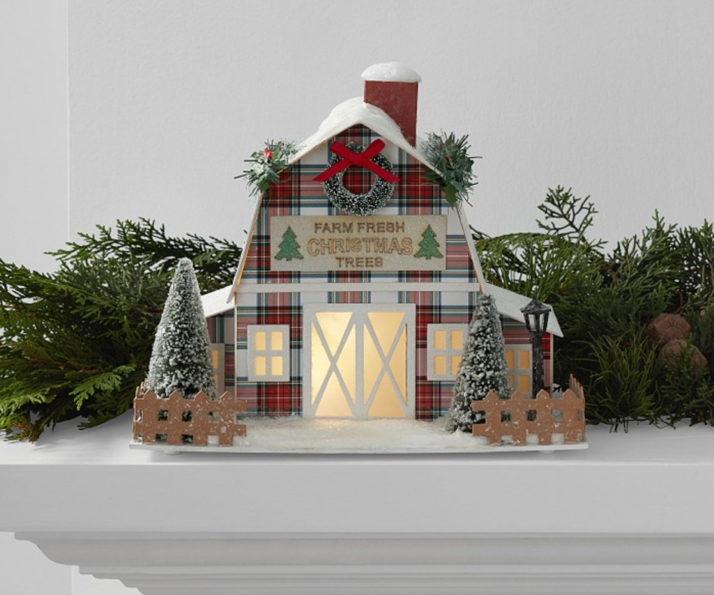 Image of Pottery Barn house that is plaid with sign that says "Farm Fresh Christmas Trees" and covered in red and green plaid. Trees and fence are in front of house with windows lit up. House is on mantel surrounded by garland.