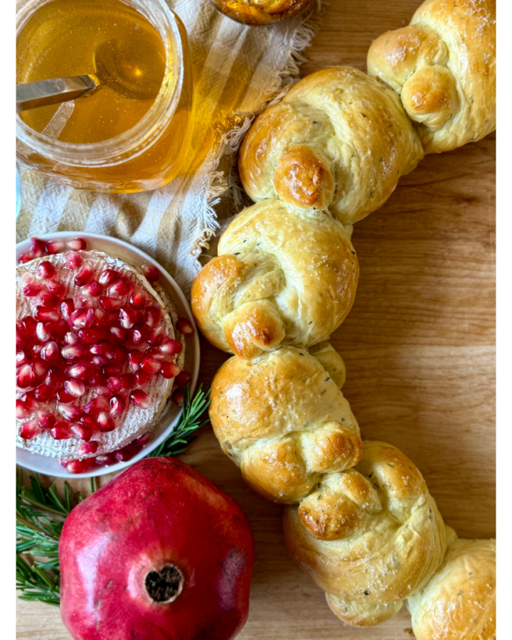 Image of half of bread wreath on wooden cutting board, surrounded by honey, brie with pomegranates, rosemary, and pomegranate.
