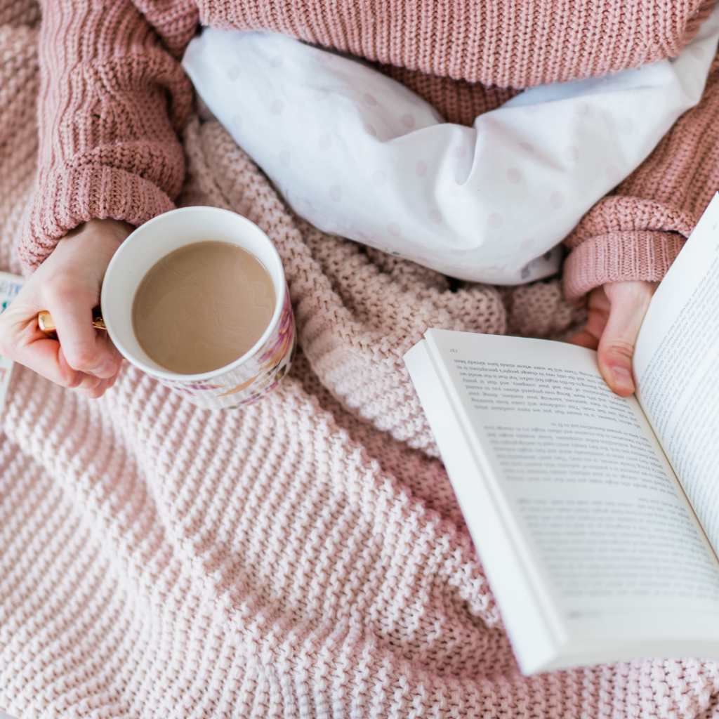 best book club books. Image of woman reading a book in bed with a cup of tea. Woman is wearing a pink knit sweater with a pink knit blanket.