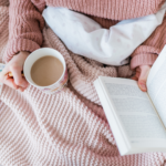 best book club books. Image of woman reading a book in bed with a cup of tea. Woman is wearing a pink knit sweater with a pink knit blanket.