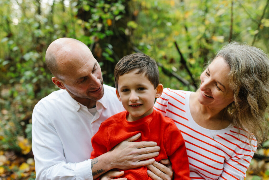 Mothering son aesthetic. Image of father, mother, and son in forest. Father is wearing a white shirt, mother is wearing a red and white stripped shirt, and son is wearing a red shirt.