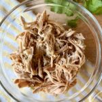 crockpot shredded chicken. Image of shredded chicken in clear glass bowl on wooden cutting board with yellow and white stripped cloth napkin, next to cilantro.