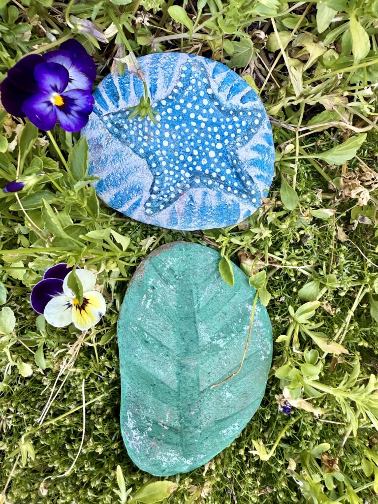 art project. Image of two painted rocks on top of each other. Rocks are painted as a star and leaf. The background is grass and flowers. 