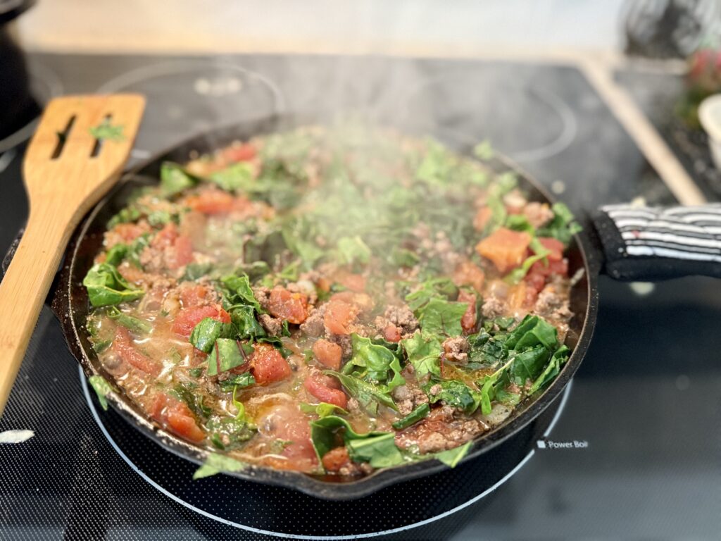 cast iron dutch oven lasagna. Image of ground beef, chard, and spinach cooking in a cast iron skillet on a stop top. 