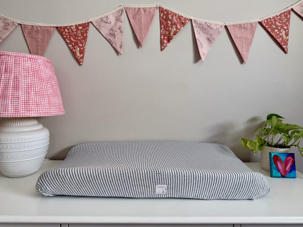 girl nursery room tour. Image of white dresser with lamp, striped changing pad, plant, and heart painting are on top to dresser. 
