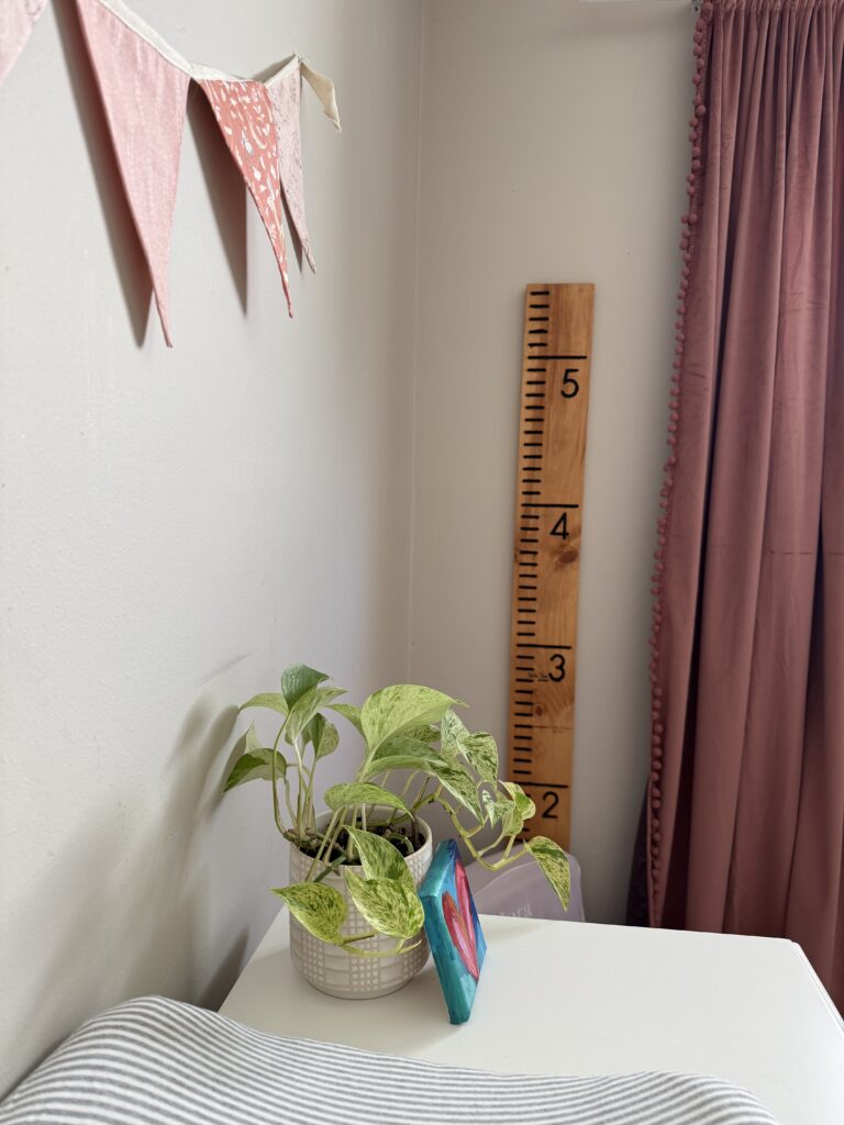 Image of diy growth chart ruler hanging on wall next to pink velvet curtains with white dresser that has plant and heart painting on canvas in front of lined changing pad.