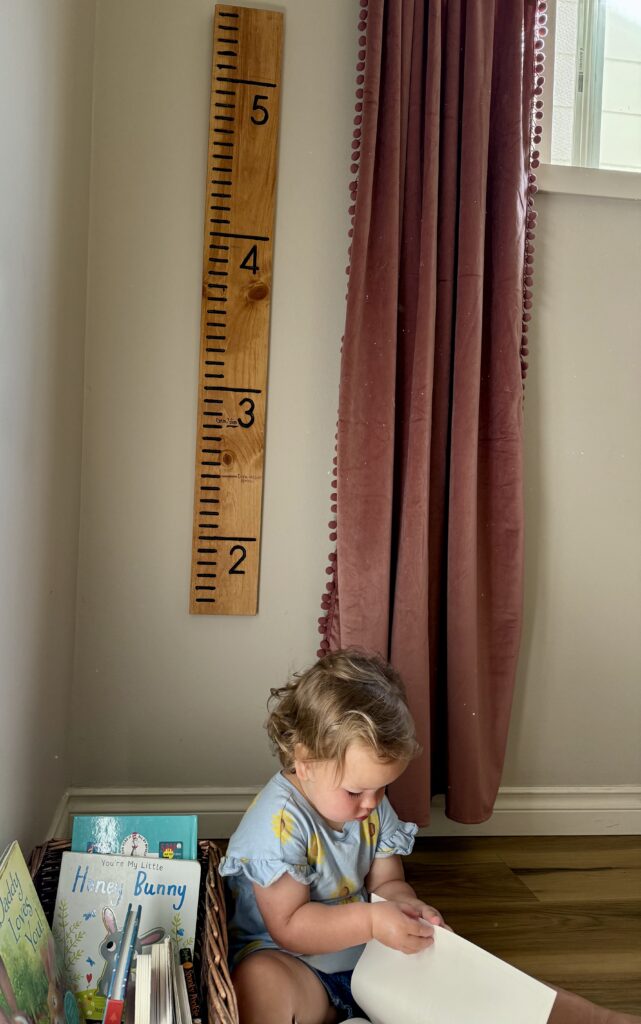 diy growth chart ruler. Image of toddler girl reading books in front of wooden growth chart ruler and pink velvet curtains.