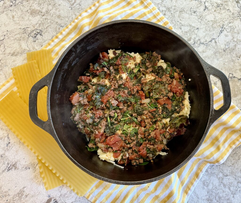 easy dinner recipes. Image of lasagna in dutch oven on a yellow and white striped towel on grey and white quartz countertop with lasagna noodles. 