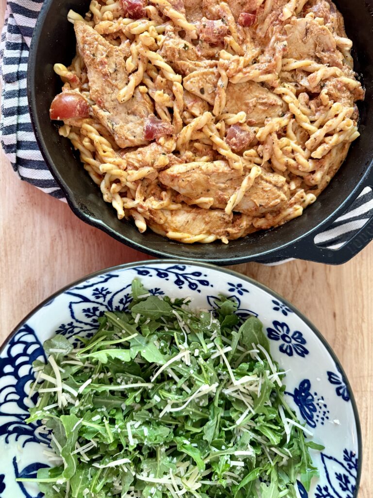 easy chicken recipe. Image of creamy bruschetta chicken in cast iron skillet on top of black and white cloth napkin on wood countertop next to arugula salad in blue and white patterned bowl.