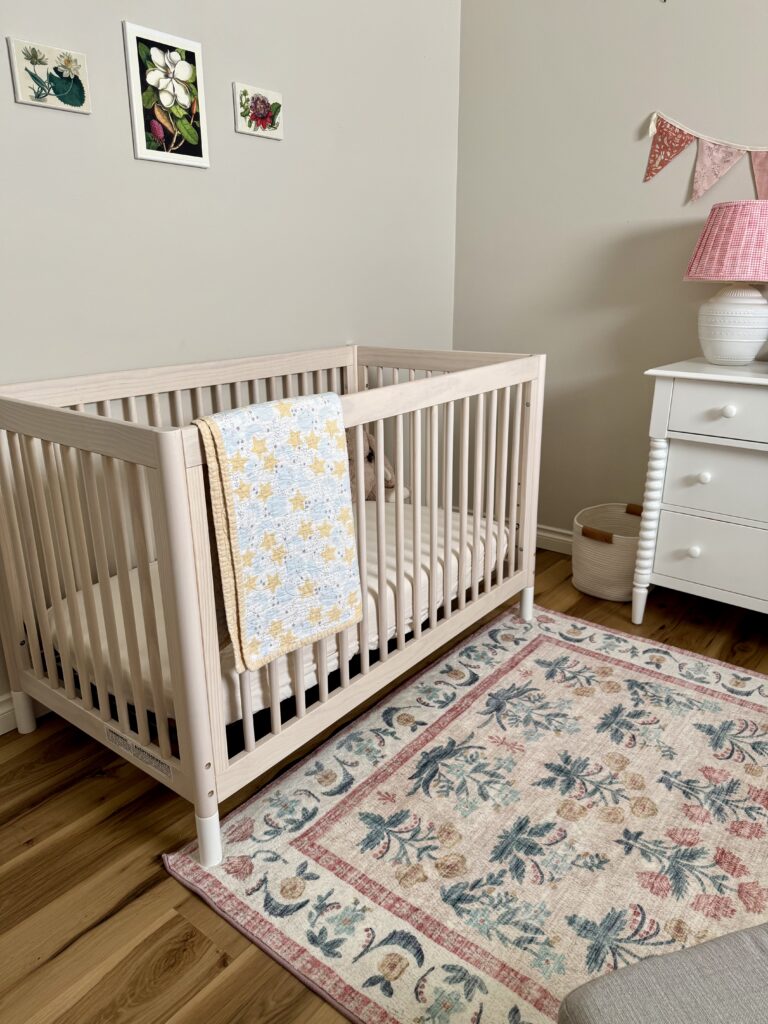 girl nursery tour. Image of crib with quilt, vintage floral art prints on wall, lamp with gingham shade, floral rug, and white dresser. 