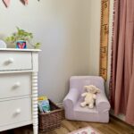how to make a growth chart ruler. image of girl's nursery with pink velvet curtains, lilac chair that says "Cora" with books in wicker basket next to it white dresser with plant and heart painting on top with fabric bunting hanging above and in front of flower print rug.