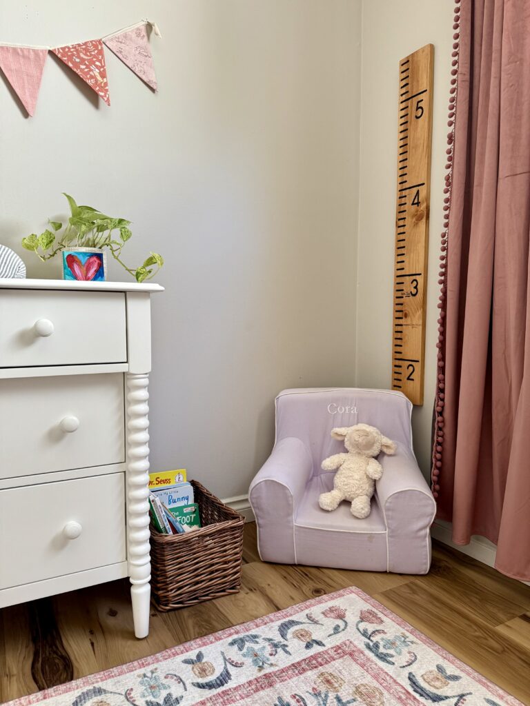 how to make a growth chart ruler. image of girl's nursery with pink velvet curtains, lilac chair that says "Cora" with books in wicker basket next to it white dresser with plant and heart painting on top with fabric bunting hanging above and in front of flower print rug.