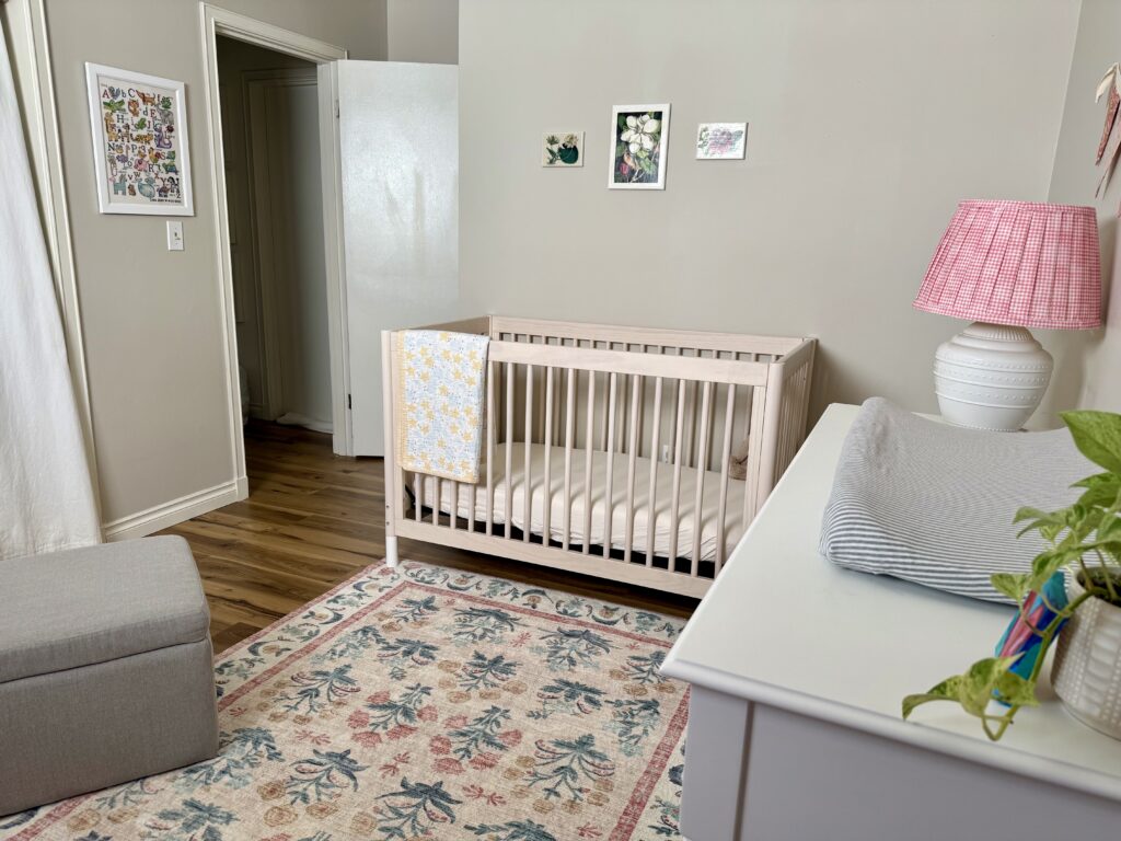 vintage inspired girls nursery. Image of crib with quilt and floral prints hanging above it. Framed cross stitch pattern hanging on the wall, dresser with lamp, striped changing pad, and plant on top. Everything is on a floral pattern rug. 