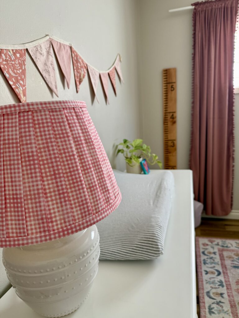 cozy baby girl nursery. Image of lamp with gingham shade, fabric bunting, striped changing pad, plant, and heart painting on top of white dresser with wooden growth chart ruler in the background, pink curtains, and floral pattern rug. 
