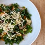easy sheet pan sausage and veggies. Image of veggies, kale, grains, and parmesan in white ceramic bowl on wood counter.