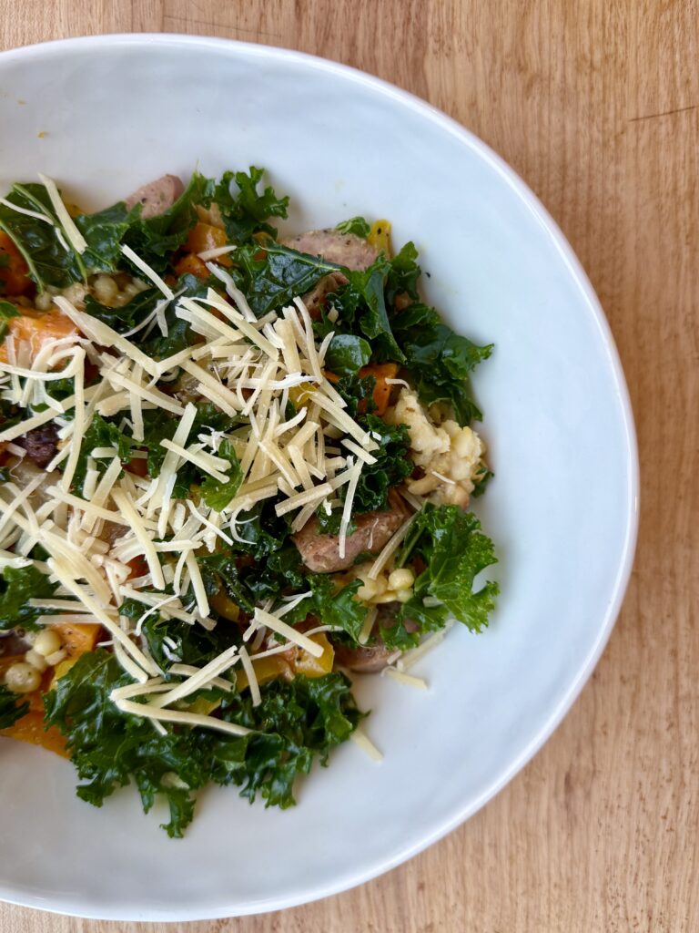 easy sheet pan sausage and veggies. Image of veggies, kale, grains, and parmesan in white ceramic bowl on wood counter.