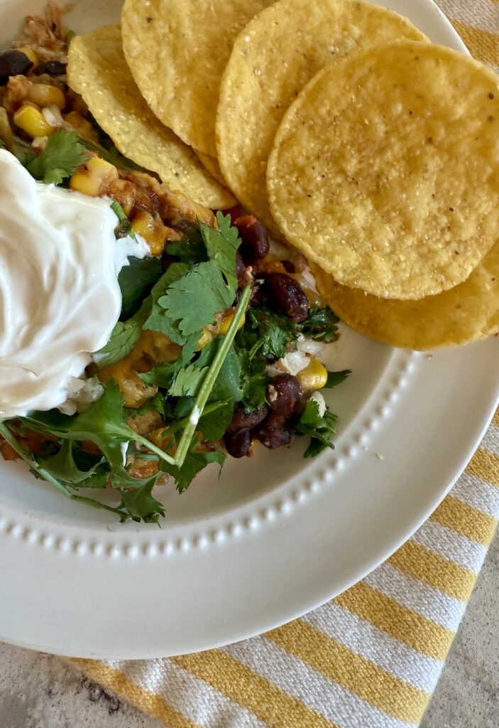 image of serving of best southwest chicken casserole topped with dollop of sour cream next to tortilla chips on white plate, sitting on yellow and white striped cloth napkin. 
