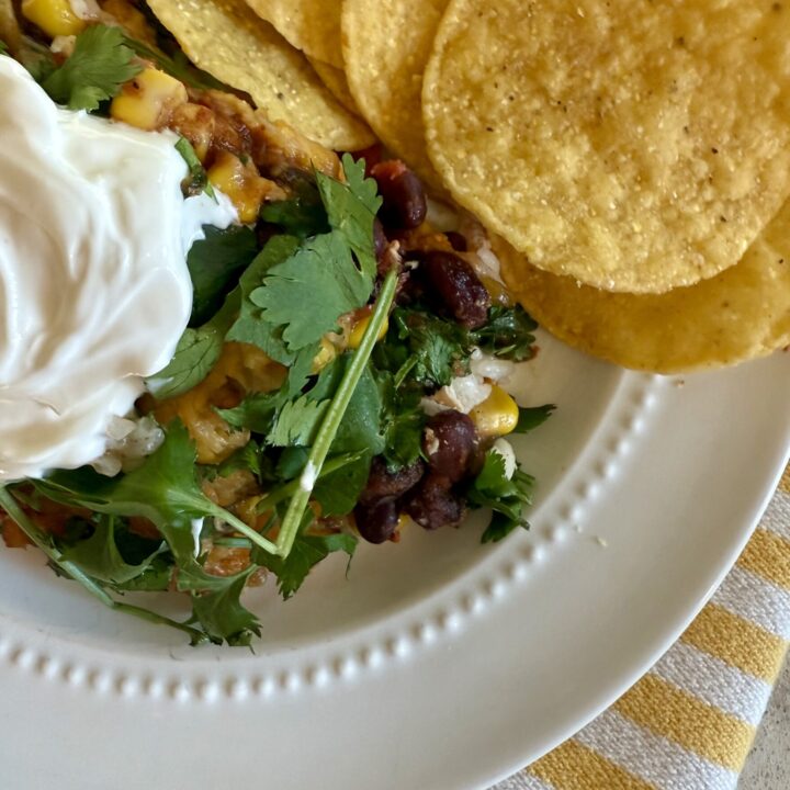 image southwest chicken casserole with sour cream on top, next to tortilla chips on a white plate, sitting on a yellow and white striped napkin.