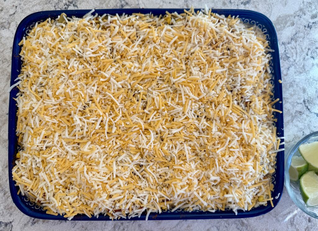 Image of healthy southwest chicken casserole topped with cheddar cheese in blue casserole dish next to bowl of sliced limes on top of white quartz countertop. 