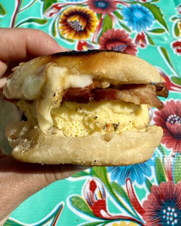 make ahead breakfast sandwiches.Image of hand holding breakfast sandwich with egg, cheese, and bacon with flower table cloth as background.