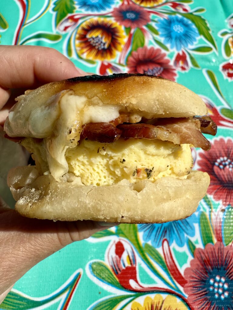 make ahead breakfast sandwiches.Image of hand holding breakfast sandwich with egg, cheese, and bacon with flower table cloth as background.