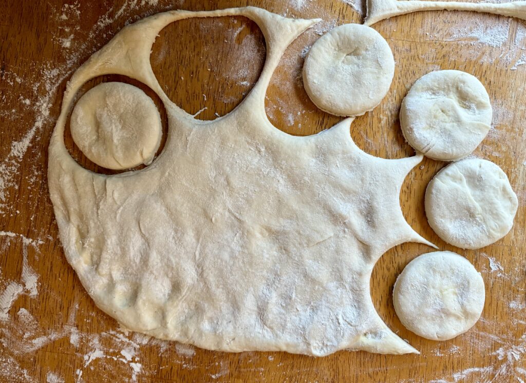 healthy make ahead breakfast sandwiches. Image of sourdough english muffin dough rolled out and english muffin cut outs on a wood counter with flour.