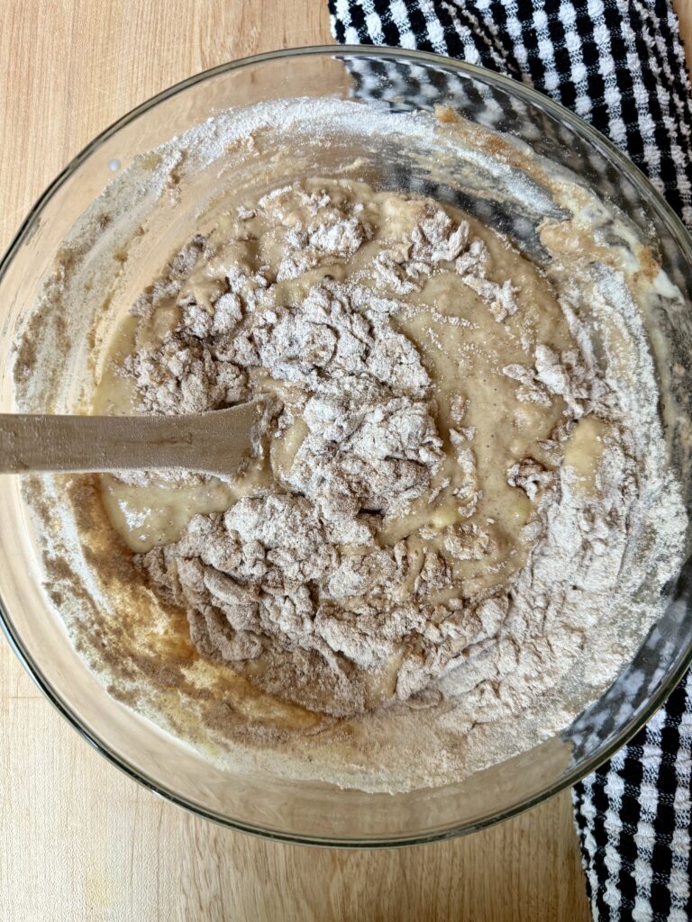 Image of wet and dry ingredients being mixed together for banana bread in glass bowl next to black and white dish cloth. 