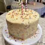 birthday traditions for kids. Image of funfetti birthday cake with rainbow sprinkles with green, pink, and purple candles on white cake platter on quartz countertop in a dining room.