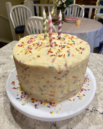birthday traditions for kids. Image of funfetti birthday cake with rainbow sprinkles with green, pink, and purple candles on white cake platter on quartz countertop in a dining room.