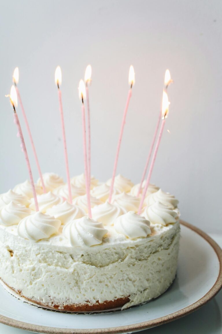 image of single layer cake with white frosting, pink lit candles on white plate with tan rim and grey background.