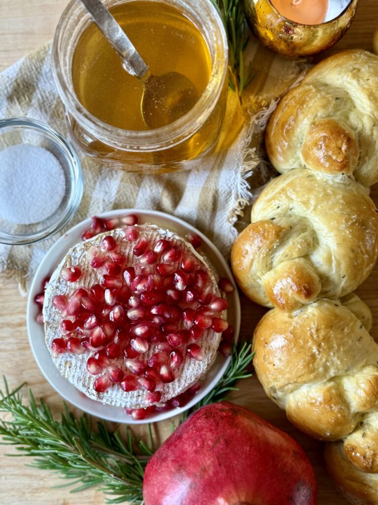 bread wreath recipe. Image of bread wreath, pomegranates on brie wheel, glass bowl of salt, glass jar of honey with spoon, sprig of rosemary, candle lit in gold votive, all on top of brown and white plaid cloth napkin. 