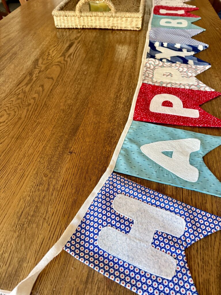 simple birthday traditions for kids. Image of bunting banner that reads "happy birthday" in blue and red pattern fabric on wood table. 