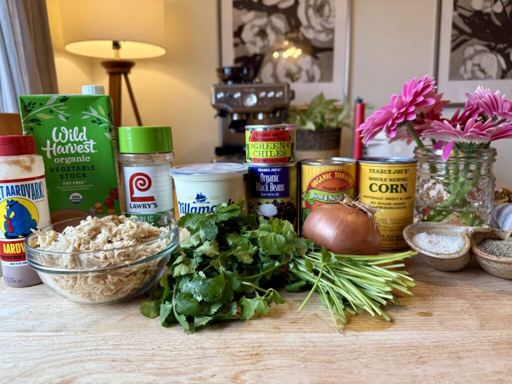 Image of ingredients for best easy chicken tortilla soup - hot sauce, shredded chicken, vegetable stock, garlic salt, cream cheese, black beans, green chilis, diced tomatoes, yellow onion, cilantro, salt and pepper with pink flowers in glass jar, a lamp in the background and two pieces of art prints of flowers. 