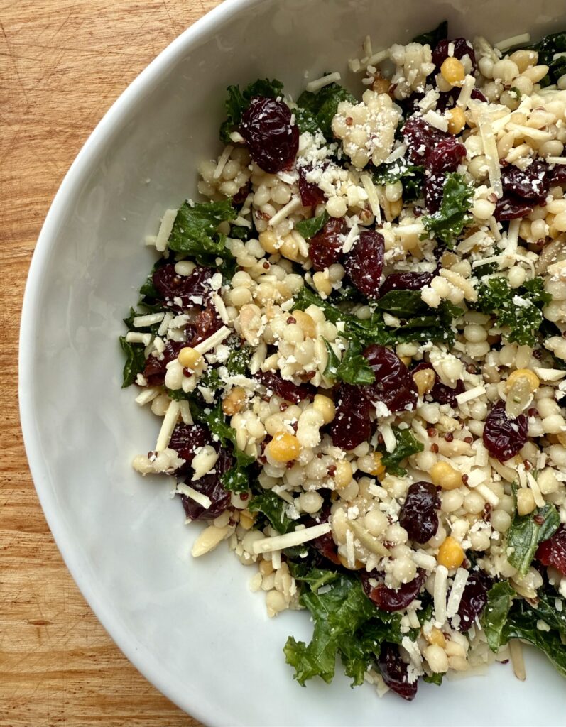 Image of easy Winter kale salad recipe. Image of bowl with kale, grains, dried cherries, and parmesan in white ceramic bowl on wooden counter top. 