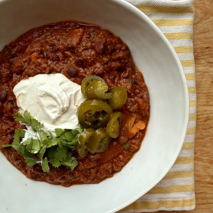 chili recipe crockpot. Image of bowl of chili with sour cream, jalapenos, and cilantro in white bowl with yellow and white striped cloth napkin on wood table.