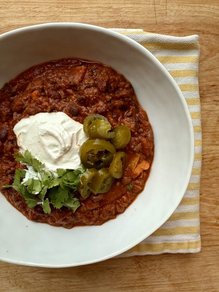 chili recipe crockpot. Image of bowl of chili with sour cream, jalapenos, and cilantro in white bowl with yellow and white striped cloth napkin on wood table.