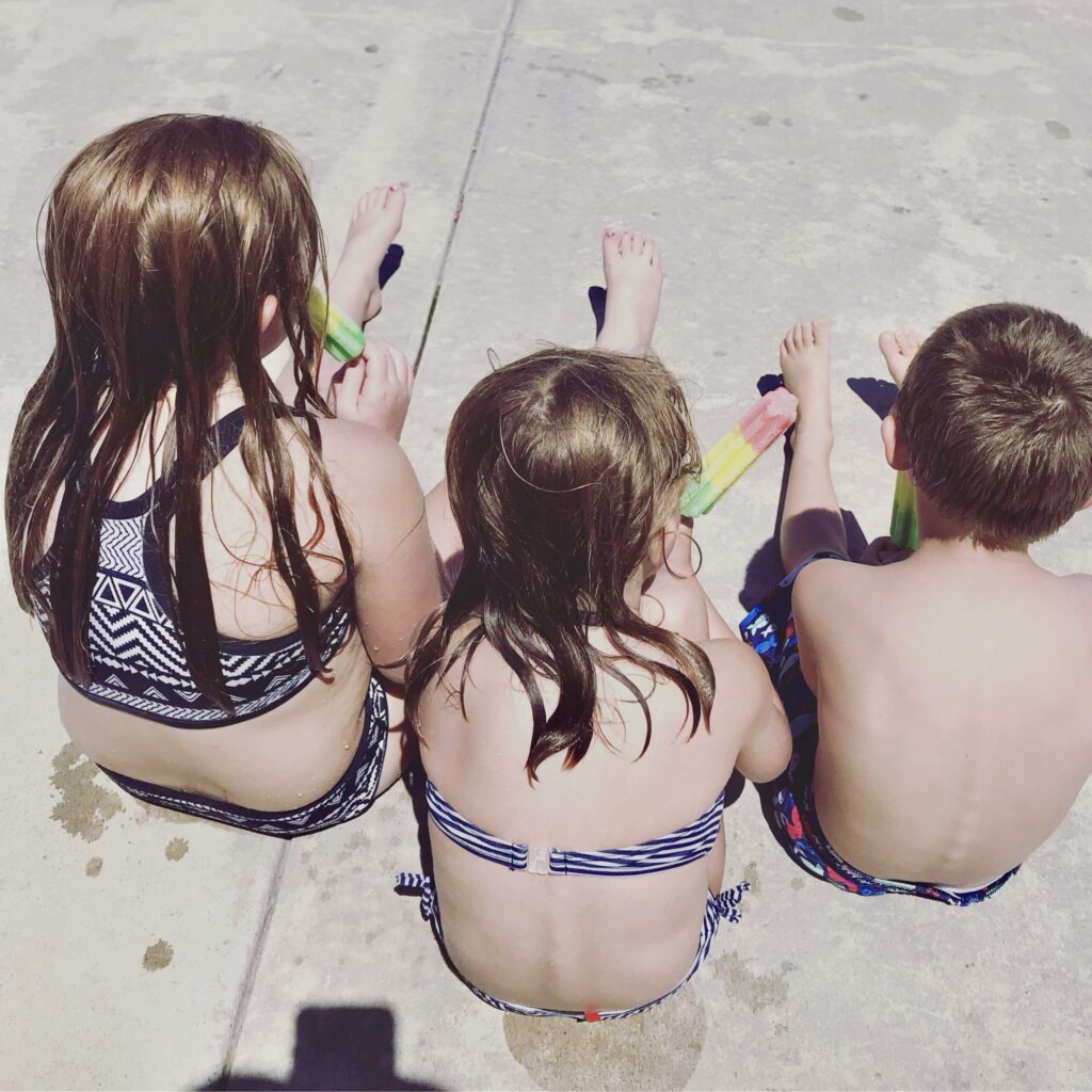 everyday motherhood. Image of 3 children in their swimsuits eating popsicles and sitting on the sidewalk.