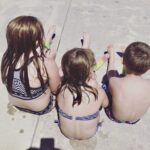 everyday motherhood. Image of 3 children in their swimsuits eating popsicles and sitting on the sidewalk.