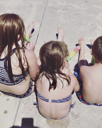 everyday motherhood. Image of 3 children in their swimsuits eating popsicles and sitting on the sidewalk.