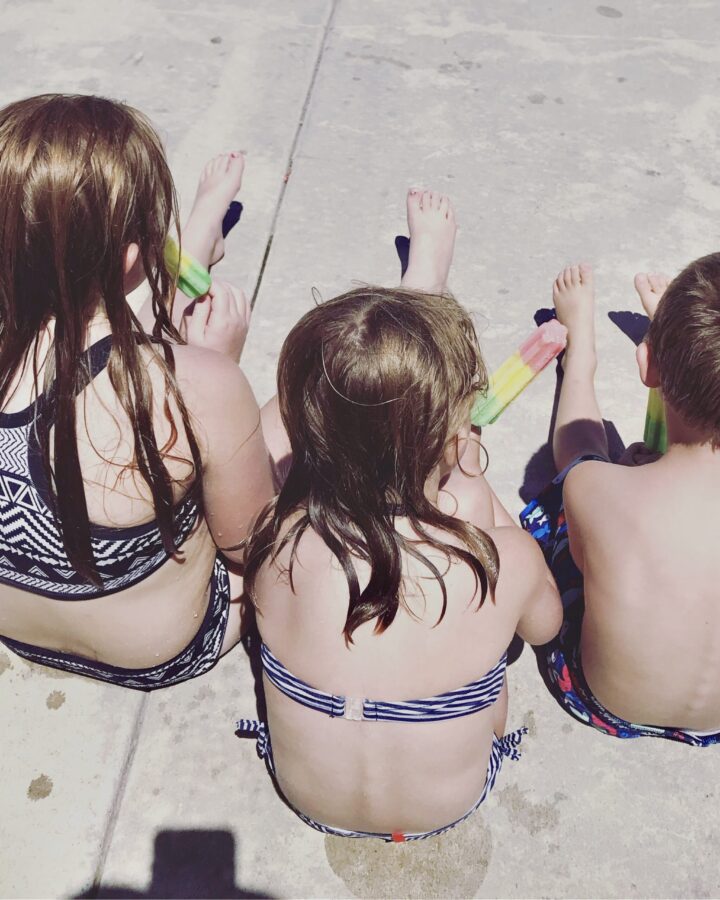 everyday motherhood. Image of 3 children in their swimsuits eating popsicles and sitting on the sidewalk.