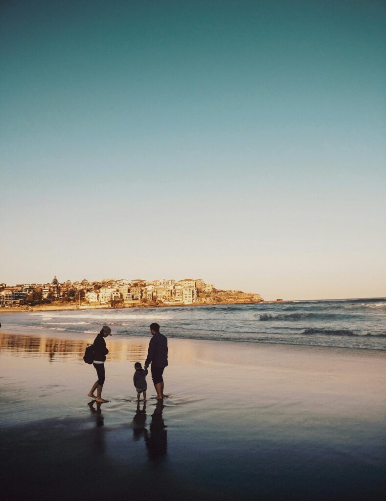 family traditions list. Image of man, woman, and child walking on beach with ocean and city in background. 