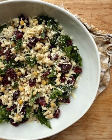 Image of kale salad recipe with kale, grains, dried cherries, and parmesan in white ceramic bowl on wooden countertop