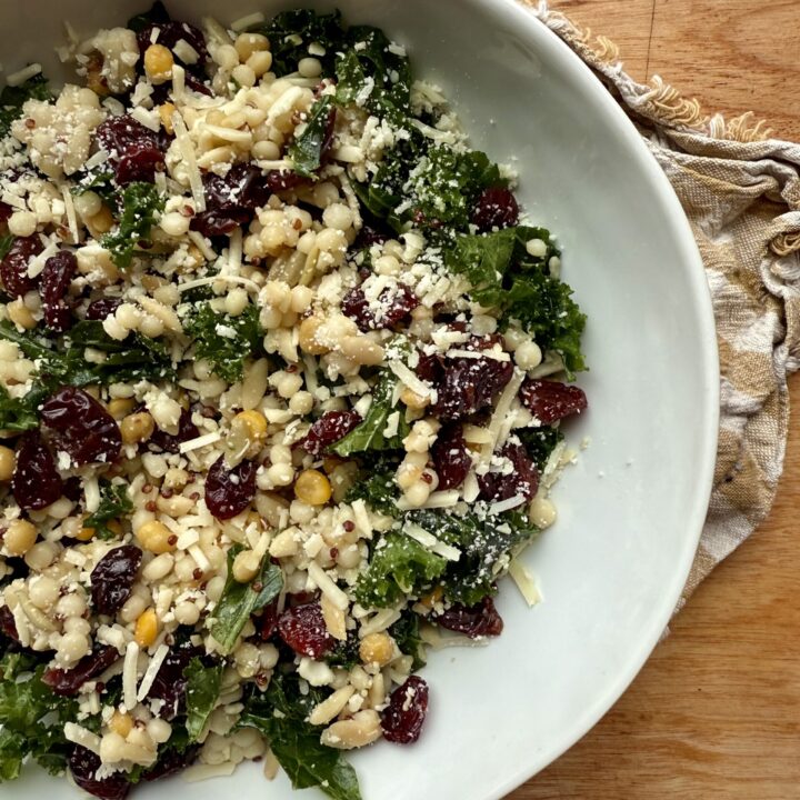 Image of kale salad recipe with kale, grains, dried cherries, and parmesan in white ceramic bowl on wooden countertop