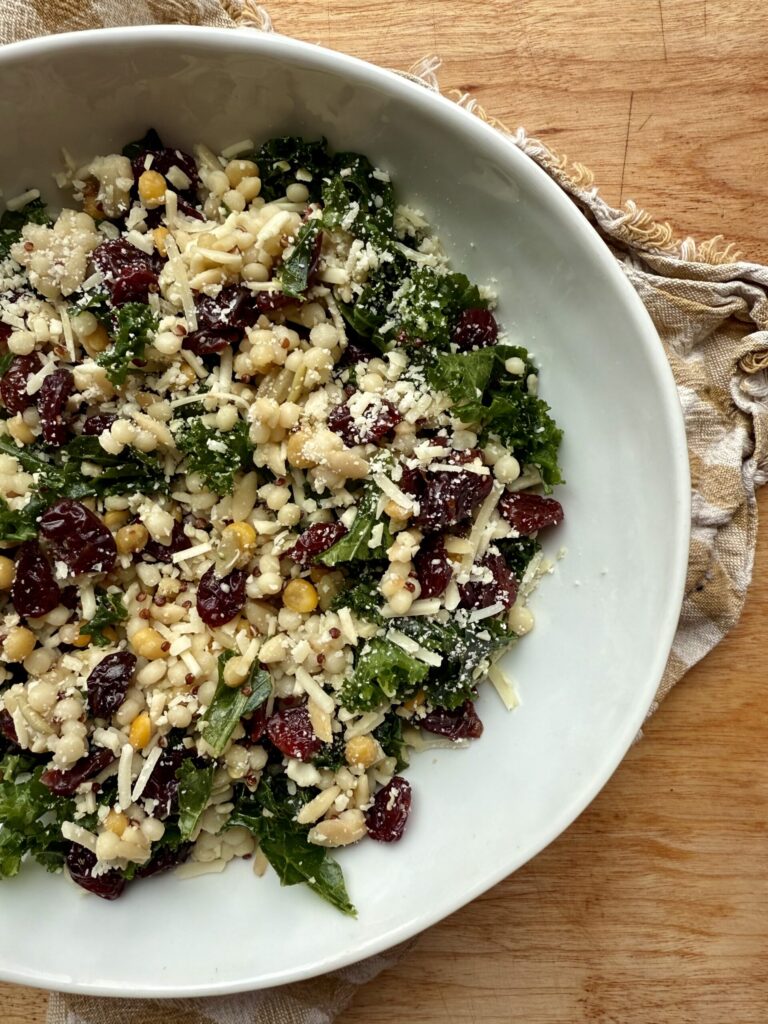 Image of kale salad recipe with kale, grains, dried cherries, and parmesan in white ceramic bowl on wooden countertop