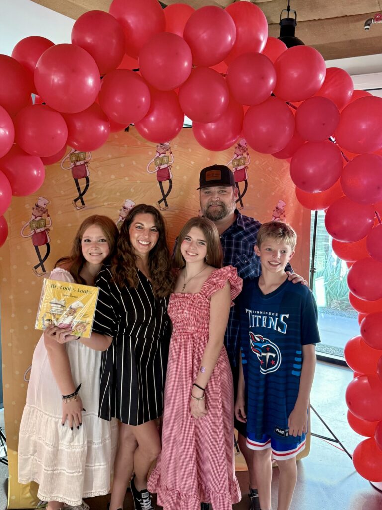 motherhood inspiration. Image of family of 5 (mom dad, two girls one boy) at a book signing under a red ballon banner.