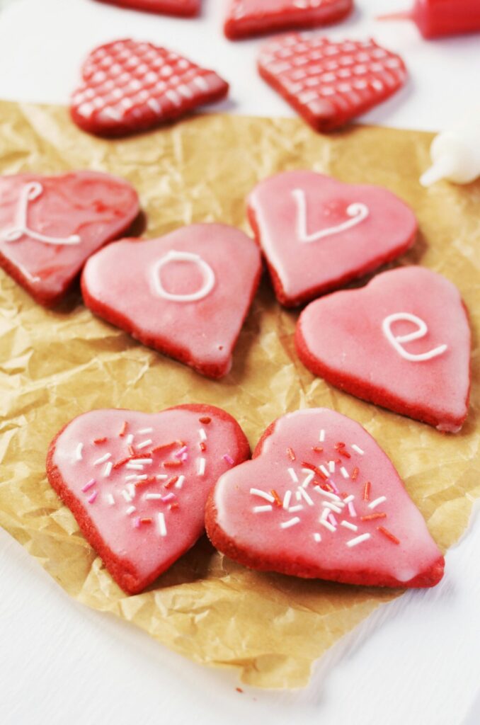 winter traditions for kids. Image of heart-shaped cookies decorated with red and white frosting and sprinkles. The cookies in the middle spell "love". The cookies are on parchment paper. 
