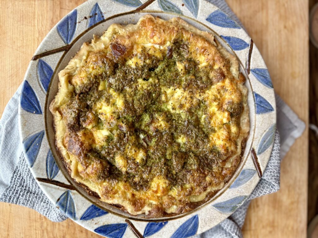 winter solstice traditions. Image of pesto quiche in pie dish with blue leaves around the edge on a wooden counter. 