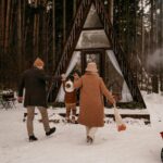 winter traditions. Image of mom and dad holding hands with toddler and walking towards A Frame cabin in snow woods.