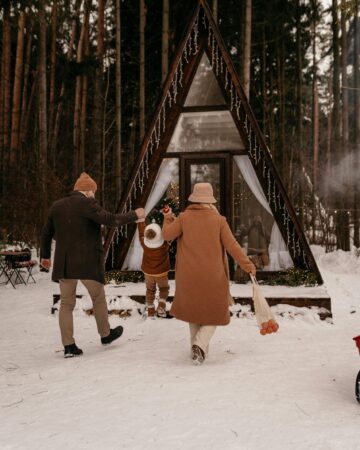 winter traditions. Image of mom and dad holding hands with toddler and walking towards A Frame cabin in snow woods.